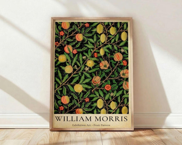 May include: Framed botanical art print featuring William Morris fruit pattern design with peaches, lemons, and berries on dark background, displayed against white wall on light wood floor. Natural wood frame contains vintage-style artwork with lush green foliage and colorful fruits in decorative Arts and Crafts movement style.