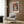 May include: Modern beige armchair with geometric patterned pillow positioned next to framed vintage-style poster showing woman wearing wide-brimmed coral hat against blue background, mounted on warm wood-paneled wall. Gold floor lamp and dark round side table with open book complete the contemporary reading nook scene with natural lighting from window.