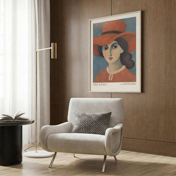 May include: Modern beige armchair with geometric patterned pillow positioned next to framed vintage-style poster showing woman wearing wide-brimmed coral hat against blue background, mounted on warm wood-paneled wall. Gold floor lamp and dark round side table with open book complete the contemporary reading nook scene with natural lighting from window.