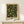 May include: Framed botanical poster with dark background featuring green foliage, orange peaches, yellow lemons, and red berries in decorative pattern. Light wood frame displays on white shelf next to small wrapped gift with twine and evergreen sprig against cream wall.