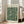 May include: Framed botanical poster with green and cream floral pattern leaning against white wall next to beige upholstered chair. Natural wood frame surrounds vintage-style William Morris design featuring intricate leaves and flowers. Bright room with large windows, hardwood floors, and natural lighting creates shadows on wall.