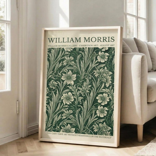 May include: Framed botanical poster with green and cream floral pattern leaning against white wall next to beige upholstered chair. Natural wood frame surrounds vintage-style William Morris design featuring intricate leaves and flowers. Bright room with large windows, hardwood floors, and natural lighting creates shadows on wall.