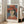 May include: Framed poster print featuring a stylized portrait of a woman wearing a wide-brimmed red hat and red top against a blue background, displayed in light wood frame leaning against white wall next to beige upholstered chair in bright, naturally-lit room with white walls and light hardwood floors.