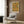 May include: Framed William Morris botanical artwork with yellow, orange, and green floral pattern displayed on wood-paneled wall above modern cream armchair with geometric pillow, brass floor lamp, and dark round side table with open book in contemporary living room setting