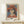 May include: Framed art print featuring a woman wearing an orange-red wide-brimmed hat and coral-colored top with white collar detail, painted in a modernist style with simplified facial features and muted color palette. Print has "PICASSO" text at bottom and sits in thin wooden frame on light wood surface next to small white wrapped gift with evergreen sprig, against cream colored wall.