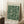 May include: Framed vintage-style poster with forest green botanical pattern featuring cream-colored flowers and flowing foliage, displayed against white paneled wall. Natural wood frame, leaning beside neutral sofa with coffee cup on wooden block nearby. Soft natural lighting creates gentle shadows on textured cream rug.