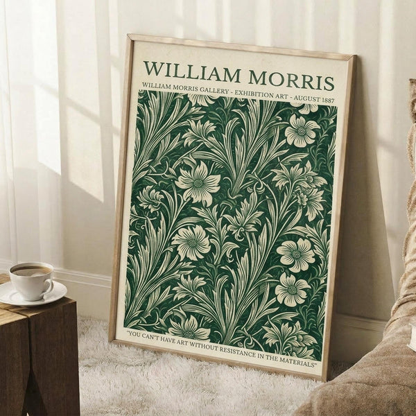 May include: Framed vintage-style poster with forest green botanical pattern featuring cream-colored flowers and flowing foliage, displayed against white paneled wall. Natural wood frame, leaning beside neutral sofa with coffee cup on wooden block nearby. Soft natural lighting creates gentle shadows on textured cream rug.