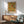 May include: Framed botanical poster with ornate floral pattern in golden yellows, orange, and green tones hanging on white wall above wooden sideboard. Mid-century modern sage green armchair with wooden frame sits beside credenza topped with white ceramic table lamp. Warm sunlight creates shadows across the minimalist interior scene.