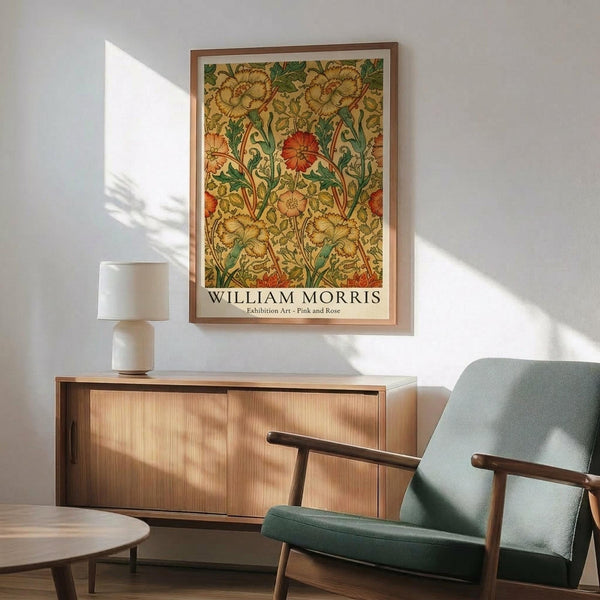 May include: Framed botanical poster with ornate floral pattern in golden yellows, orange, and green tones hanging on white wall above wooden sideboard. Mid-century modern sage green armchair with wooden frame sits beside credenza topped with white ceramic table lamp. Warm sunlight creates shadows across the minimalist interior scene.