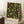 May include: Framed botanical poster with black background featuring colorful fruit and foliage pattern in green, yellow, orange and red tones, leaning against white wall next to white coffee cup on wooden surface and beige fabric.