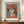 May include: Framed poster print featuring a stylized portrait of a woman wearing an orange-red wide-brimmed hat and matching top with white collar ties. The artwork shows geometric facial features and dark hair against a blue-gray background, labeled "PICASSO" at bottom. The brown wooden frame sits on a walnut credenza beside a cream ceramic vase and white books in an elegant room with white paneled walls.