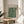 May include: Framed William Morris exhibition poster with ornate green and cream floral pattern displayed on light wood round side table next to beige upholstered chair, with plant leaves visible and natural sunlight creating shadows on white wall behind