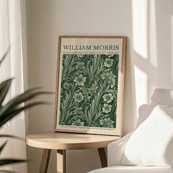 May include: Framed William Morris exhibition poster with ornate green and cream floral pattern displayed on light wood round side table next to beige upholstered chair, with plant leaves visible and natural sunlight creating shadows on white wall behind
