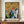 May include: Framed exhibition poster featuring William Morris floral wallpaper pattern with ornate yellow and orange blooms, green foliage, and modern abstract geometric shapes in blues and greens below. Brown wooden frame displayed on walnut credenza beside white ceramic vase and stacked books in cream-colored room with white wainscoting.