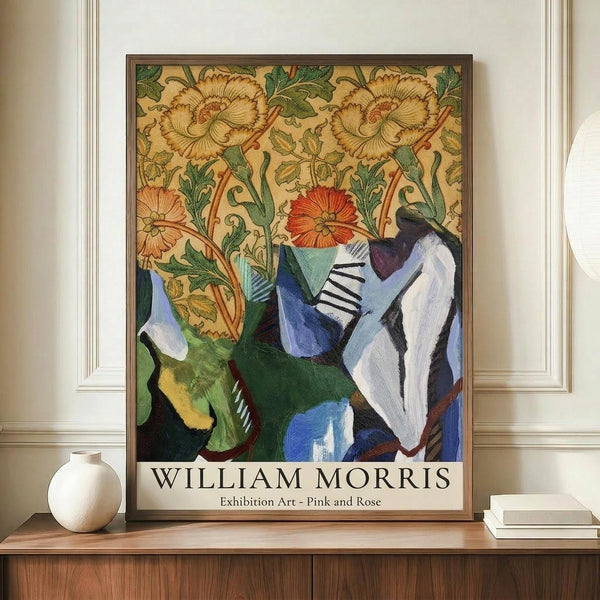May include: Framed exhibition poster featuring William Morris floral wallpaper pattern with ornate yellow and orange blooms, green foliage, and modern abstract geometric shapes in blues and greens below. Brown wooden frame displayed on walnut credenza beside white ceramic vase and stacked books in cream-colored room with white wainscoting.