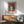 May include: Framed art print of woman wearing orange-red wide-brimmed hat and matching top against blue background, displayed above mid-century modern wooden credenza with white ceramic lamp. Mint green upholstered armchair with wooden frame sits nearby on wooden floor. Natural sunlight creates geometric shadows on white wall, giving warm ambiance to contemporary living room setting.