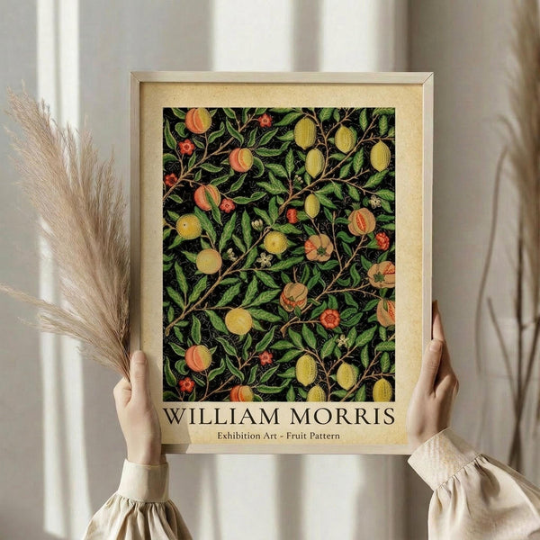 May include: Person in cream blouse holding framed botanical art print featuring William Morris-style fruit and foliage pattern with oranges, lemons, and green leaves on dark background. Natural wood frame with cream matting. Dried pampas grass stems visible beside frame against soft white curtained window backdrop.