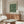 May include: Framed botanical poster with green floral pattern displayed above wooden credenza in modern living room. White curved chair and glass oval coffee table visible. Poster shows intricate leaf and flower design in dark green tones with cream text header. Natural lighting creates shadows across warm wood furniture and neutral walls.