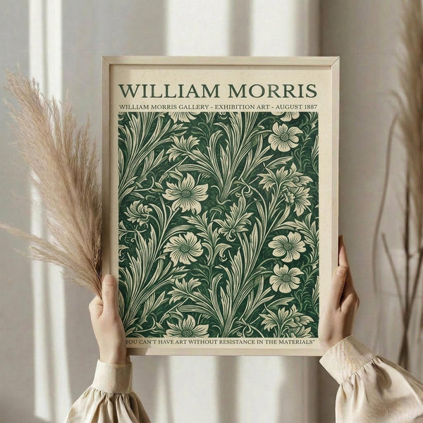 May include: Person holding framed vintage-style poster featuring William Morris floral pattern in deep green with cream botanical motifs. Light wood frame against white curtained background. Dried pampas grass visible on left side. Cream-colored long sleeve top with gathered cuffs shown.