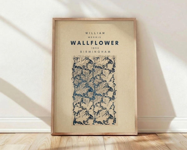 May include: Framed vintage botanical wallpaper design print in cream and blue tones, featuring ornate floral and vine patterns in Arts and Crafts style. Natural wood frame leaning against white wall on light wooden floor with soft shadows from window light.