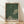 May include: Framed poster print leaning against white wall on light wood floor. Green background with white polka dots creating optical illusion pattern. Two cream-colored circular sections with black dots form abstract shapes. Bold black text reads "YAYOI KUSAMA" with Japanese characters and "TOKYO" below. Natural wood frame with visible grain. Soft shadows cast on textured white wall from natural lighting.