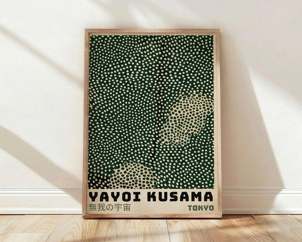 May include: Framed poster print leaning against white wall on light wood floor. Green background with white polka dots creating optical illusion pattern. Two cream-colored circular sections with black dots form abstract shapes. Bold black text reads "YAYOI KUSAMA" with Japanese characters and "TOKYO" below. Natural wood frame with visible grain. Soft shadows cast on textured white wall from natural lighting.