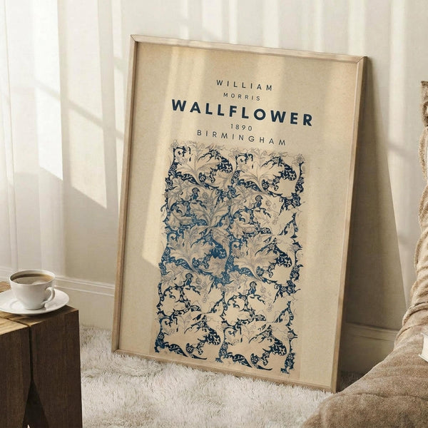 May include: Framed vintage botanical poster with blue and beige floral pattern leaning against white wall corner. Light wood frame surrounds cream-colored print featuring intricate wallflower design. Cozy room setting with white coffee cup on wooden block, soft beige throw blanket, and natural window light creating gentle shadows on textured rug.