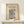 May include: Framed vintage-style poster with William Morris wallflower pattern in blue and cream tones, displayed on light wood console table against cream wall. Small white wrapped gift with greenery sits nearby. Natural lighting creates soft shadows on the warm, minimalist setting.