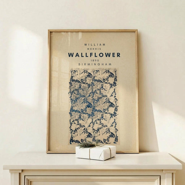 May include: Framed vintage-style poster with William Morris wallflower pattern in blue and cream tones, displayed on light wood console table against cream wall. Small white wrapped gift with greenery sits nearby. Natural lighting creates soft shadows on the warm, minimalist setting.