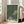 May include: Framed poster with green background covered in white polka dots, featuring two large cream-colored circular shapes with smaller dark dots inside. Text reads "YAYOI KUSAMA" in bold black letters with Japanese characters below and "TOKYO" in smaller text. Light wood frame leaning against white wall next to beige upholstered sofa in bright, sunlit room with hardwood floors.