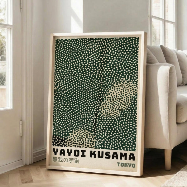 May include: Framed poster with green background covered in white polka dots, featuring two large cream-colored circular shapes with smaller dark dots inside. Text reads "YAYOI KUSAMA" in bold black letters with Japanese characters below and "TOKYO" in smaller text. Light wood frame leaning against white wall next to beige upholstered sofa in bright, sunlit room with hardwood floors.