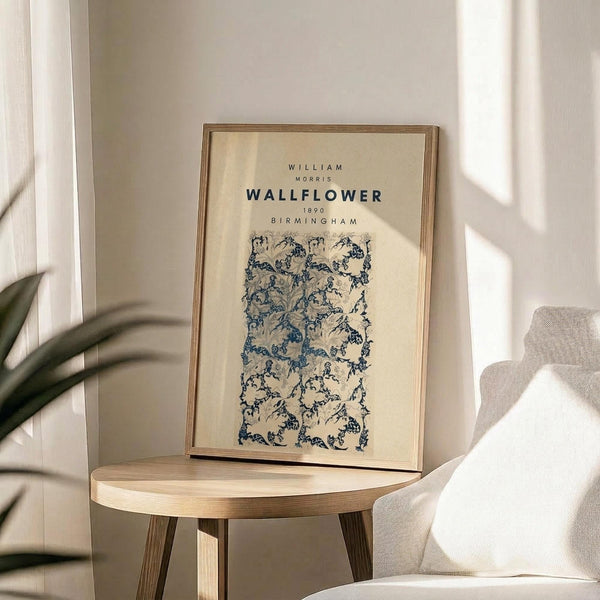 May include: Framed poster with cream background featuring blue botanical wallpaper pattern displayed on light wood round side table next to beige upholstered chair. Natural sunlight streams across white wall creating geometric shadows. Green plant leaves visible in corner. Poster shows intricate floral design in navy blue on off-white background within thin wooden frame.