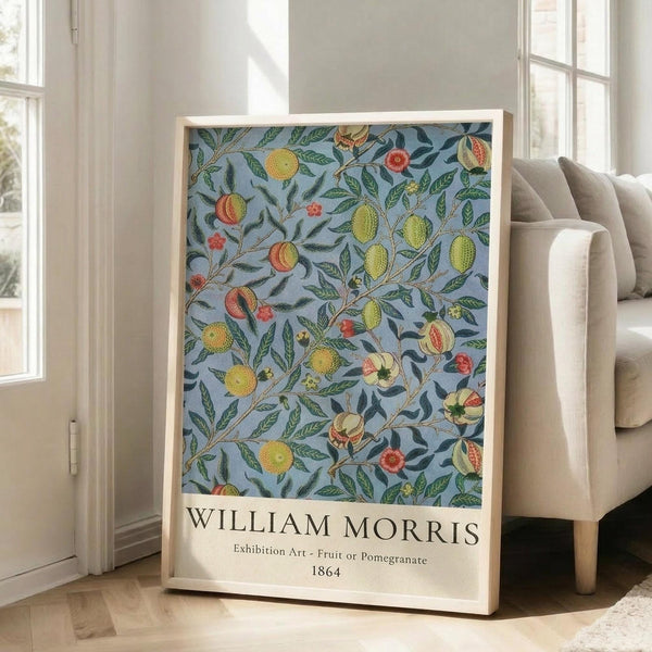 May include: Large framed botanical art print leaning against white wall beside beige upholstered chair. The artwork features William Morris's decorative pattern with fruits, pomegranates, and foliage in yellows, oranges, and greens on light blue background. Natural wood frame, sunlit room with tall windows and hardwood floors.