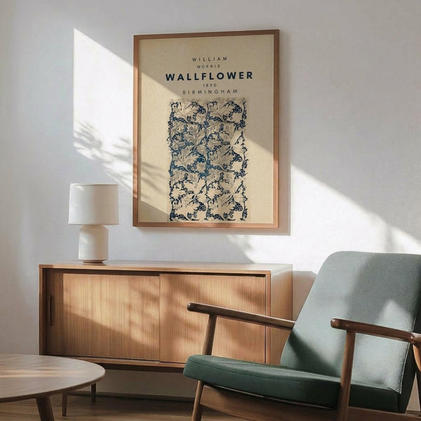 May include: Framed William Morris botanical wallpaper design print in blue and cream tones hanging on white wall above wooden sideboard. Mid-century modern room setting with sage green upholstered chair, small round side table, and white ceramic table lamp. Natural sunlight creates geometric shadows across the scene. The vintage floral pattern features intricate climbing vines and flowers in a repeating design within a light wood frame.