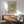 May include: Vintage William Morris botanical poster with green floral pattern displayed above wooden sideboard in mid-century modern living room. Sage green upholstered armchair with wooden frame sits nearby, white ceramic table lamp on credenza. Natural sunlight creates shadows on white wall, showing warm wood tones throughout furniture pieces.