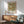 May include: Framed William Morris exhibition poster featuring botanical trellis pattern with blue birds, red roses, and green foliage on cream background, displayed above mid-century modern wooden credenza with white ceramic lamp, next to sage green upholstered armchair with wooden arms in sunlit room with white walls