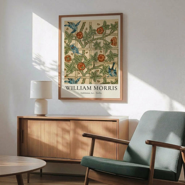 May include: Framed William Morris exhibition poster featuring botanical trellis pattern with blue birds, red roses, and green foliage on cream background, displayed above mid-century modern wooden credenza with white ceramic lamp, next to sage green upholstered armchair with wooden arms in sunlit room with white walls