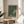 May include: Framed art print with dark green background covered in white polka dots, featuring two lighter circular areas creating an infinity-like pattern. Text reads "YAYOI KUSAMA" and "TOKYO" in black letters at bottom. Light wood frame sits on round wooden side table next to beige upholstered chair. Plant leaves visible on left, white walls with natural sunlight streaming across scene.