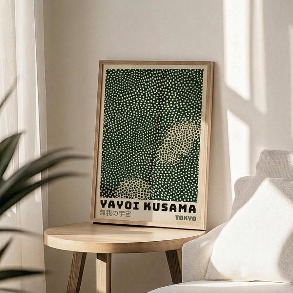 May include: Framed art print with dark green background covered in white polka dots, featuring two lighter circular areas creating an infinity-like pattern. Text reads "YAYOI KUSAMA" and "TOKYO" in black letters at bottom. Light wood frame sits on round wooden side table next to beige upholstered chair. Plant leaves visible on left, white walls with natural sunlight streaming across scene.
