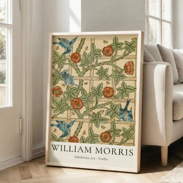 May include: Framed botanical poster with William Morris Trellis pattern featuring blue birds, orange flowers, and green vines on cream background with lattice design. Light wood frame leans against white wall next to beige upholstered chair in bright room with large windows and natural sunlight streaming across wooden floors.