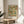 May include: Framed exhibition poster featuring William Morris's Trellis pattern with green vines, orange roses, and blue birds on cream background with wooden lattice design. Natural wood frame displayed on round wooden side table next to beige upholstered chair. Soft natural lighting creates shadows across neutral wall with plant visible in corner.