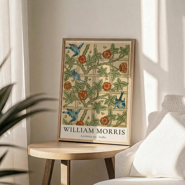May include: Framed exhibition poster featuring William Morris's Trellis pattern with green vines, orange roses, and blue birds on cream background with wooden lattice design. Natural wood frame displayed on round wooden side table next to beige upholstered chair. Soft natural lighting creates shadows across neutral wall with plant visible in corner.