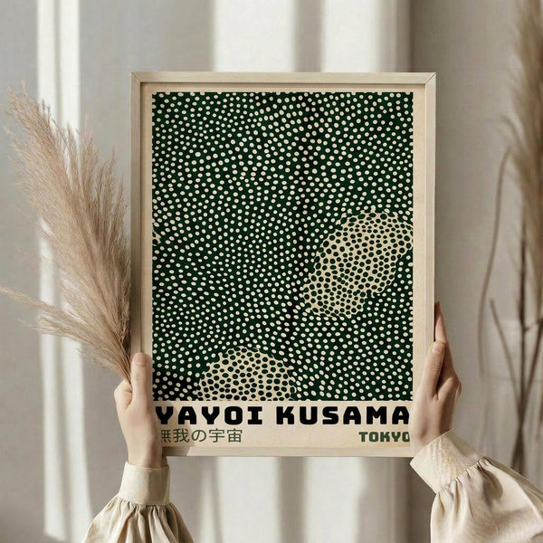 May include: Person in cream sweater holding framed art print featuring dark green background with white polka dots and cream colored circular shapes creating abstract pattern. Black text at bottom reads "YAYOI KUSAMA" with Japanese characters and "TOKYO". Light wood frame against neutral wall with dried pampas grass visible on left side.