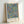 May include: Framed art print leaning against white wall showing William Morris botanical pattern with fruits and foliage on light blue background. Natural wood frame contains colorful design featuring pomegranates, citrus fruits, green leaves and small red flowers in vintage Arts and Crafts style. Exhibition poster format with artist name and artwork title at bottom.