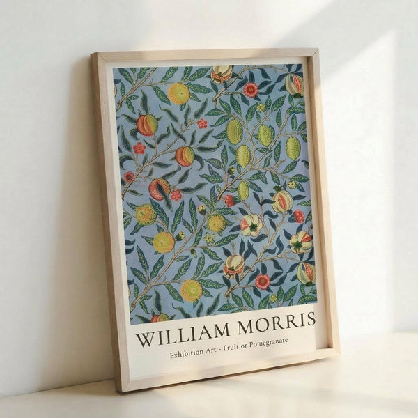 May include: Framed art print leaning against white wall showing William Morris botanical pattern with fruits and foliage on light blue background. Natural wood frame contains colorful design featuring pomegranates, citrus fruits, green leaves and small red flowers in vintage Arts and Crafts style. Exhibition poster format with artist name and artwork title at bottom.