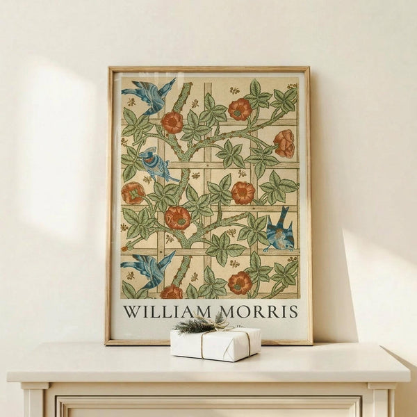 May include: Framed botanical poster featuring William Morris's Trellis pattern with blue birds perched among orange roses and green foliage on a cream background with geometric grid lines. Light wood frame displayed on white wooden furniture beside a small wrapped gift with twine and evergreen sprig against neutral wall.