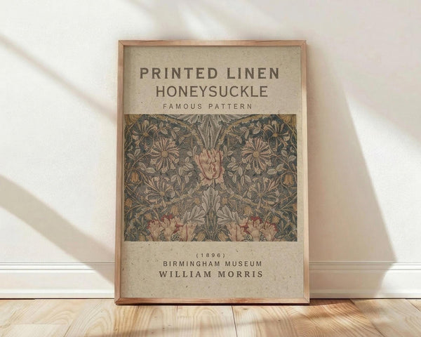 May include: Framed poster print featuring William Morris's Honeysuckle pattern from 1896, displayed against white wall on light wood floor. Vintage botanical design shows intricate pink and coral flowers with gray-green foliage in Arts and Crafts style. Light wood frame contains beige background with pattern sample and museum attribution text. Natural window lighting creates soft shadows on textured white wall.