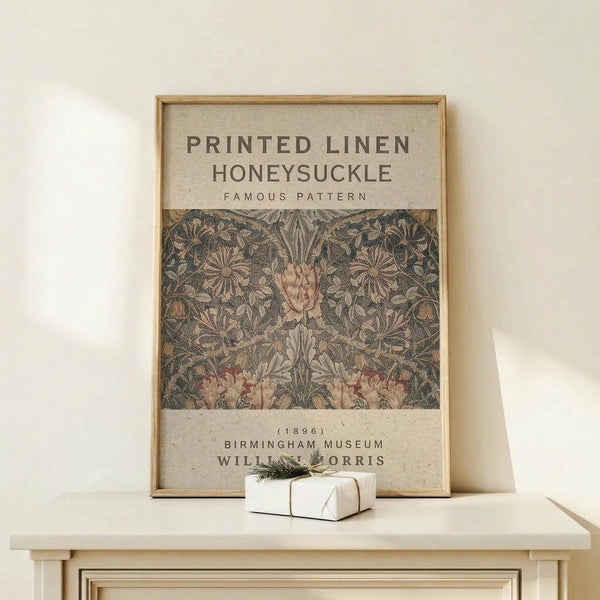 May include: Framed vintage botanical poster featuring William Morris honeysuckle pattern in muted green and pink tones, displayed on light wood console table against cream wall. Small white gift box with greenery sits beside the natural wood frame. The artwork shows intricate floral design with text indicating it's from Birmingham Museum, dated 1896.