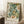 May include: Framed botanical art print leaning against white wall corner, featuring large cream and peach flowers with lush green foliage in William Morris style. Light wood frame on white textured rug beside small wooden stool with white coffee cup. Soft natural lighting creates geometric shadows on wall. Cozy beige throw blanket visible at right edge.