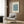 May include: Framed poster with blue floral pattern and "MARIGOLD" and "WILLIAM MORRIS" text displayed on wood-paneled wall above beige upholstered chair with geometric patterned pillow. Modern brass floor lamp with white base stands nearby next to dark round side table with open book. Neutral toned room setting with natural lighting from window.